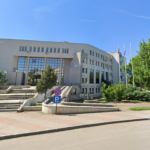 Beige municipal building with large glass entrance, steps, and flagpoles, set against a blue sky and surrounding greenery.