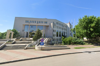 Beige municipal building with large glass entrance, steps, and flagpoles, set against a blue sky and surrounding greenery.