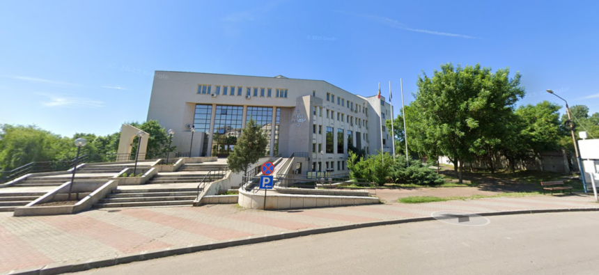 Beige municipal building with large glass entrance, steps, and flagpoles, set against a blue sky and surrounding greenery.