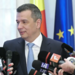 Man in a dark suit and light blue tie speaks to reporters, surrounded by multiple microphones; flags in the background suggest a formal press conference.