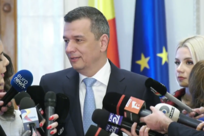 Man in a dark suit and light blue tie speaks to reporters, surrounded by multiple microphones; flags in the background suggest a formal press conference.