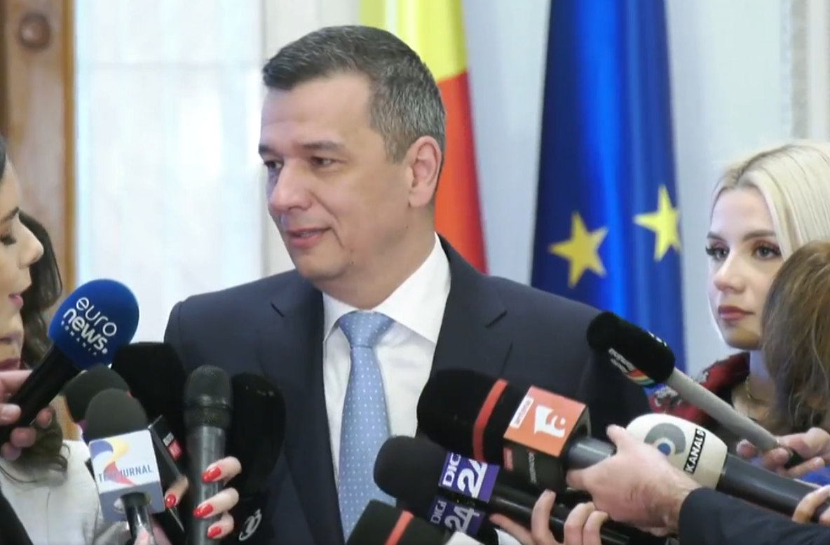 Man in a dark suit and light blue tie speaks to reporters, surrounded by multiple microphones; flags in the background suggest a formal press conference.