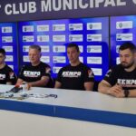 Four men in black KEMPO shirts sit at a press conference table with microphones, against a blue backdrop covered with logos.