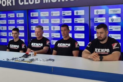 Four men in black KEMPO shirts sit at a press conference table with microphones, against a blue backdrop covered with logos.