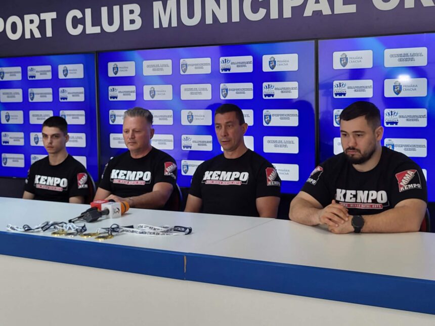 Four men in black KEMPO shirts sit at a press conference table with microphones, against a blue backdrop covered with logos.