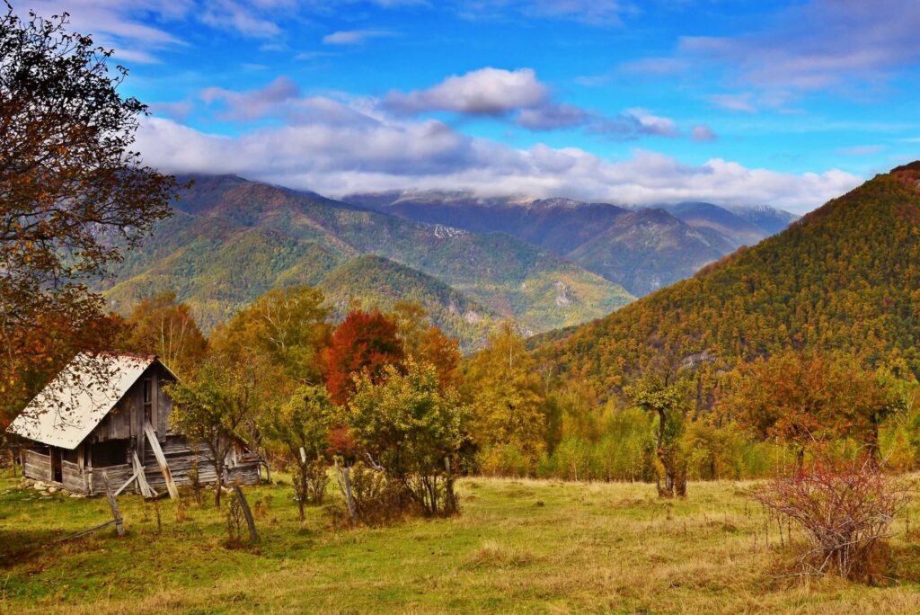Autumn mountain landscape with a weathered wooden cabin among colorful trees and distant hills under a blue sky.