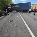Highway collision scene with a red truck and a blue trailer, debris scattered on the road and bystanders nearby against a blue sky and trees left of the scene.