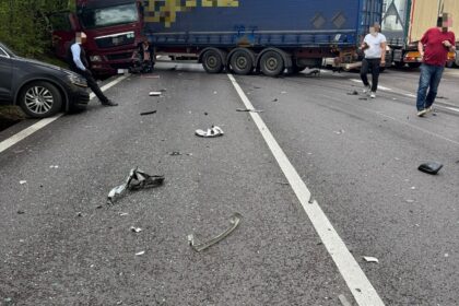Highway collision scene with a red truck and a blue trailer, debris scattered on the road and bystanders nearby against a blue sky and trees left of the scene.