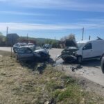 Two cars severely damaged in a head-on collision on a rural road, with debris and trucks nearby and emergency responders on the scene.