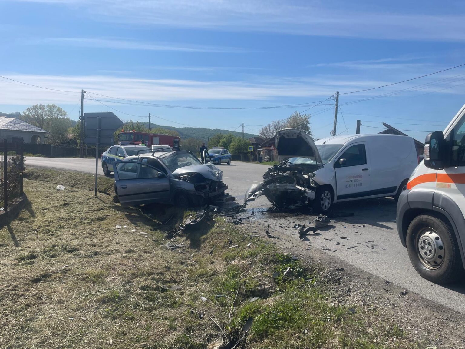 Two cars severely damaged in a head-on collision on a rural road, with debris and trucks nearby and emergency responders on the scene.