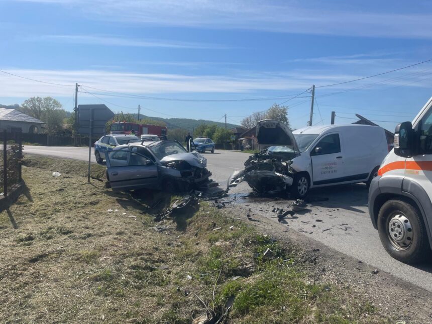 Two cars severely damaged in a head-on collision on a rural road, with debris and trucks nearby and emergency responders on the scene.