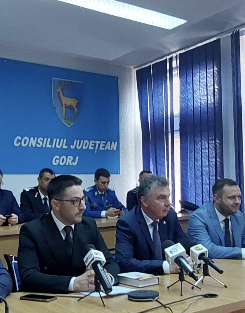 Press conference at the Gorj County Council chamber; men in suits sit at a long table with microphones, one reading a book, others ready to speak, blue council wall with crest behind them.
