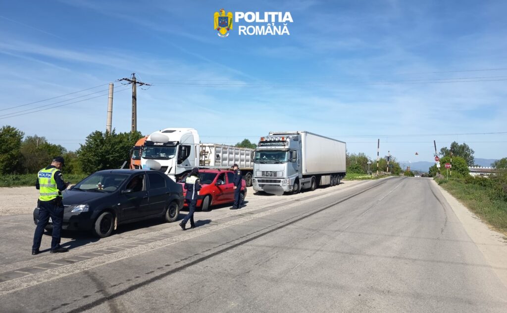 Romanian police officers conduct a roadside checkpoint, inspecting a black car while a red car is stopped beside a white truck on a rural road.
