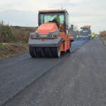 Road construction scene with an orange asphalt roller smoothing fresh pavement; workers in the distance.