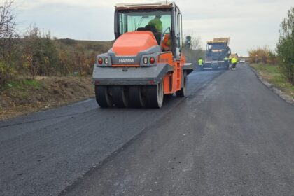 Road construction scene with an orange asphalt roller smoothing fresh pavement; workers in the distance.
