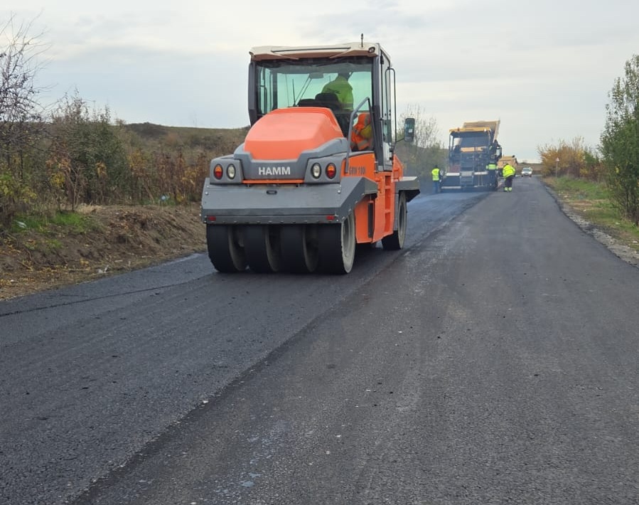 Road construction scene with an orange asphalt roller smoothing fresh pavement; workers in the distance.