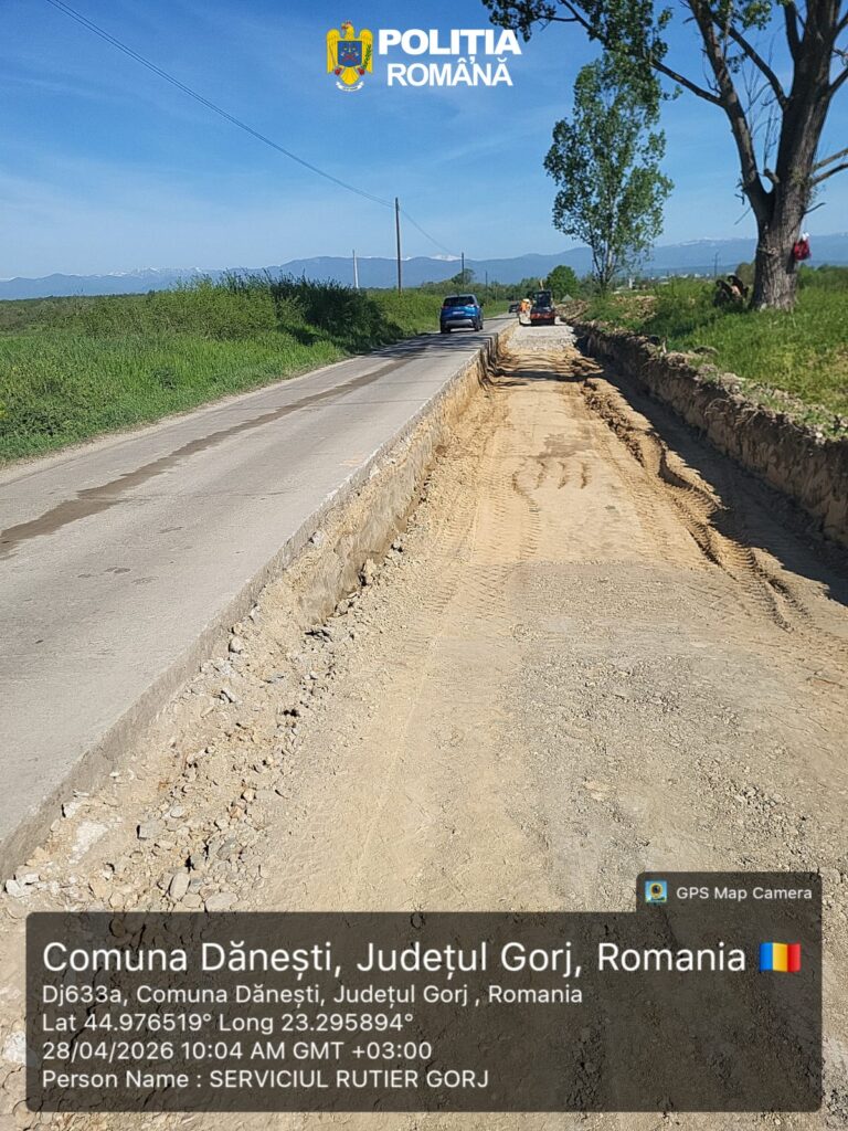 Rural road under construction in Dănești, Gorj, Romania, with a wide dirt trench on the right, two cars driving ahead, green fields and distant mountains under a blue sky. Police Romania watermark at the top.