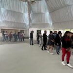 People lined up indoors in a white tent with a draped ceiling, waiting near a long table setup.