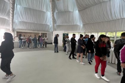 People lined up indoors in a white tent with a draped ceiling, waiting near a long table setup.