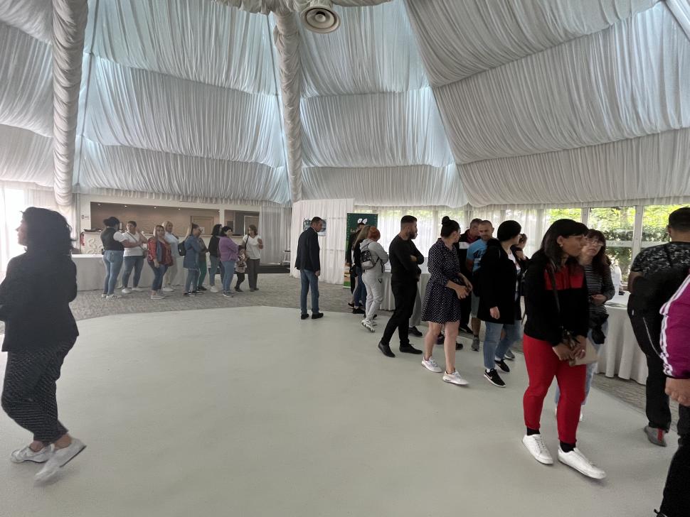 People lined up indoors in a white tent with a draped ceiling, waiting near a long table setup.