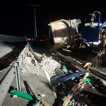 Nighttime highway crash scene with a toppled white dump truck trailer and scattered debris along a damaged guardrail area