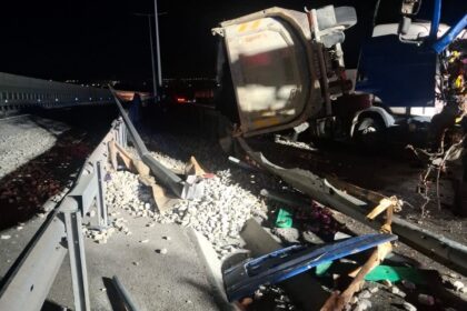 Nighttime highway crash scene with a toppled white dump truck trailer and scattered debris along a damaged guardrail area