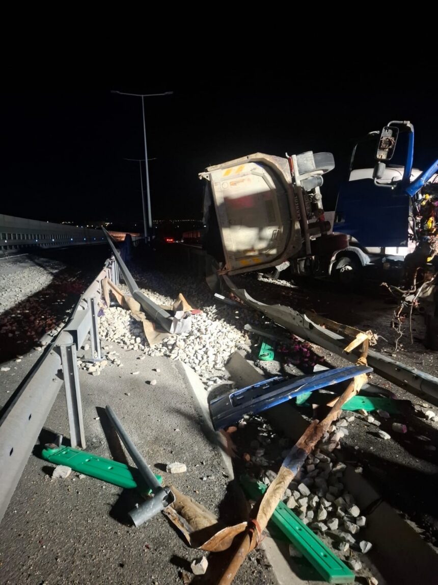 Nighttime highway crash scene with a toppled white dump truck trailer and scattered debris along a damaged guardrail area