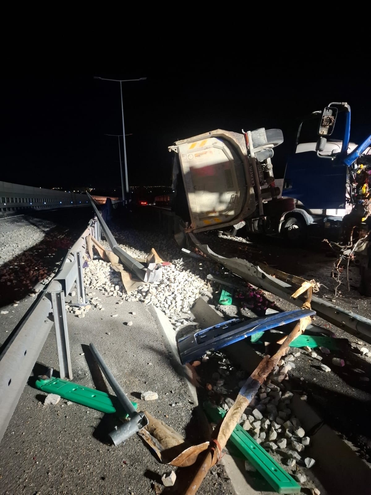 Nighttime highway crash scene with a toppled white dump truck trailer and scattered debris along a damaged guardrail area