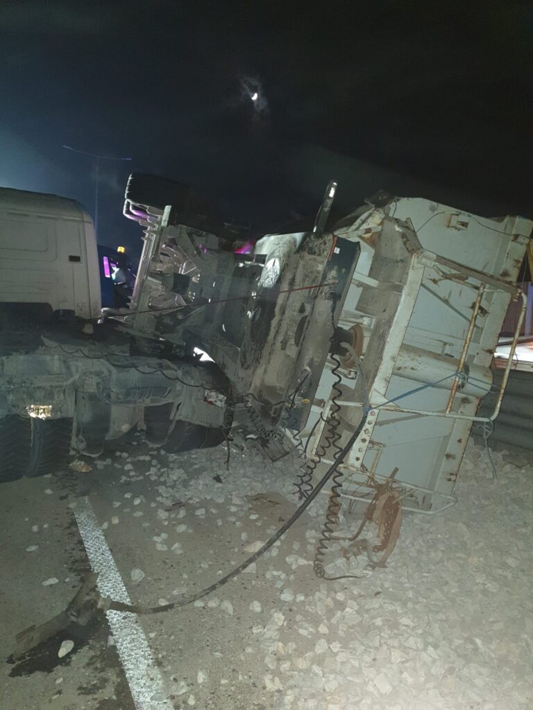 Nighttime highway crash: a damaged truck trailer toppled on its side amid debris and scattered rocks/ice on the road.