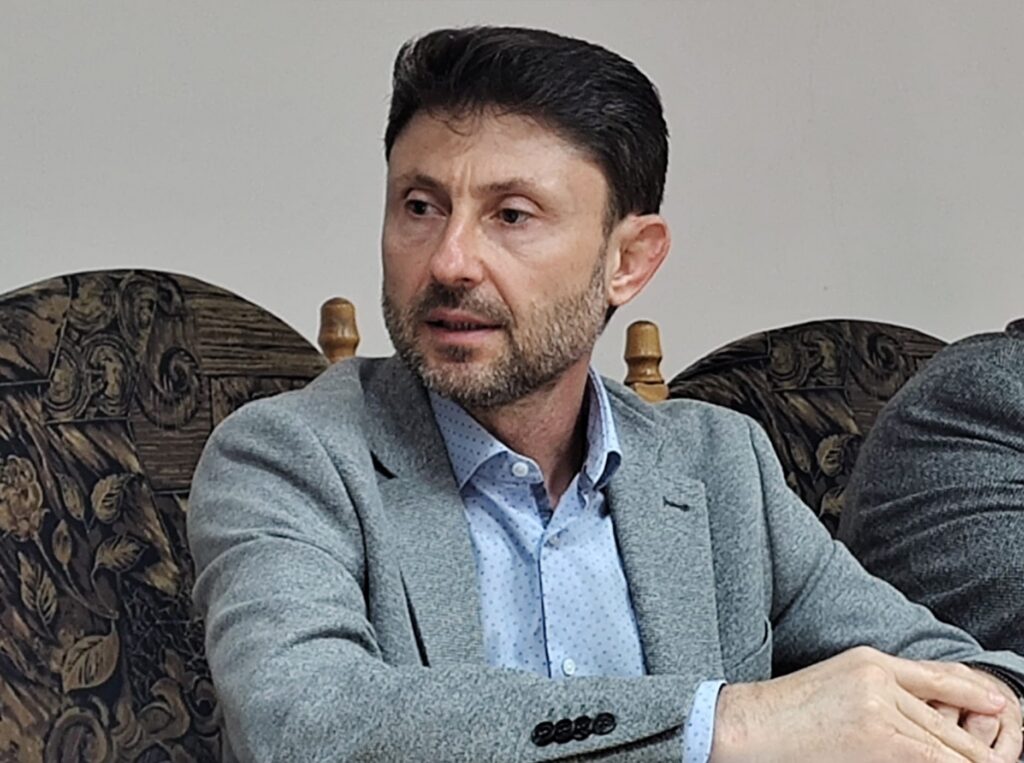 Man in a gray blazer and light blue shirt seated at a conference table, looking to his left.