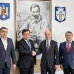 Four men in business suits stand in a formal room; two in the center shake hands while the others stand nearby, with a large framed portrait and crest emblems on the wall behind them.