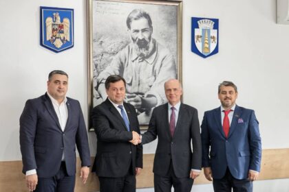 Four men in business suits stand in a formal room; two in the center shake hands while the others stand nearby, with a large framed portrait and crest emblems on the wall behind them.