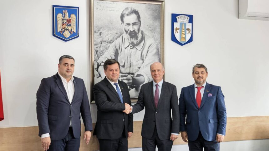 Four men in business suits stand in a formal room; two in the center shake hands while the others stand nearby, with a large framed portrait and crest emblems on the wall behind them.