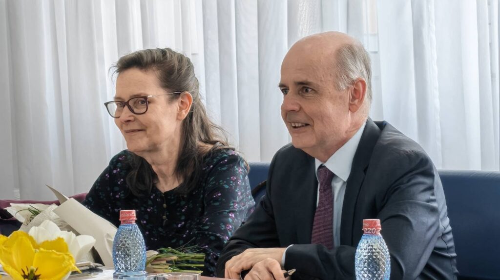 Two adults sit at a conference table with flowers, papers, and water bottles, smiling during a meeting.