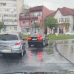 Blurred urban street after rain: cars drive on a wet, reflective road with puddles and blurred buildings in the background.