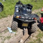 Rear view of a muddy ATV stuck in a deep rut, with a trailer hitch and rear rack in a grassy, dirt area.