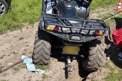 Rear view of a muddy ATV stuck in a deep rut, with a trailer hitch and rear rack in a grassy, dirt area.