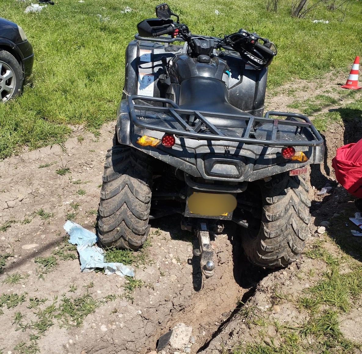 Rear view of a muddy ATV stuck in a deep rut, with a trailer hitch and rear rack in a grassy, dirt area.