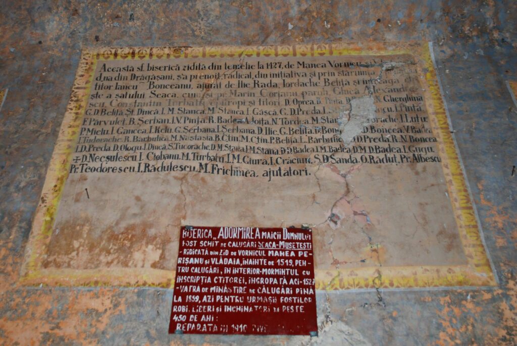 Weathered wall with a large beige panel of dense Romanian text, framed by a yellow border and rust-streaked surroundings, plus a red plaque below describing more details.