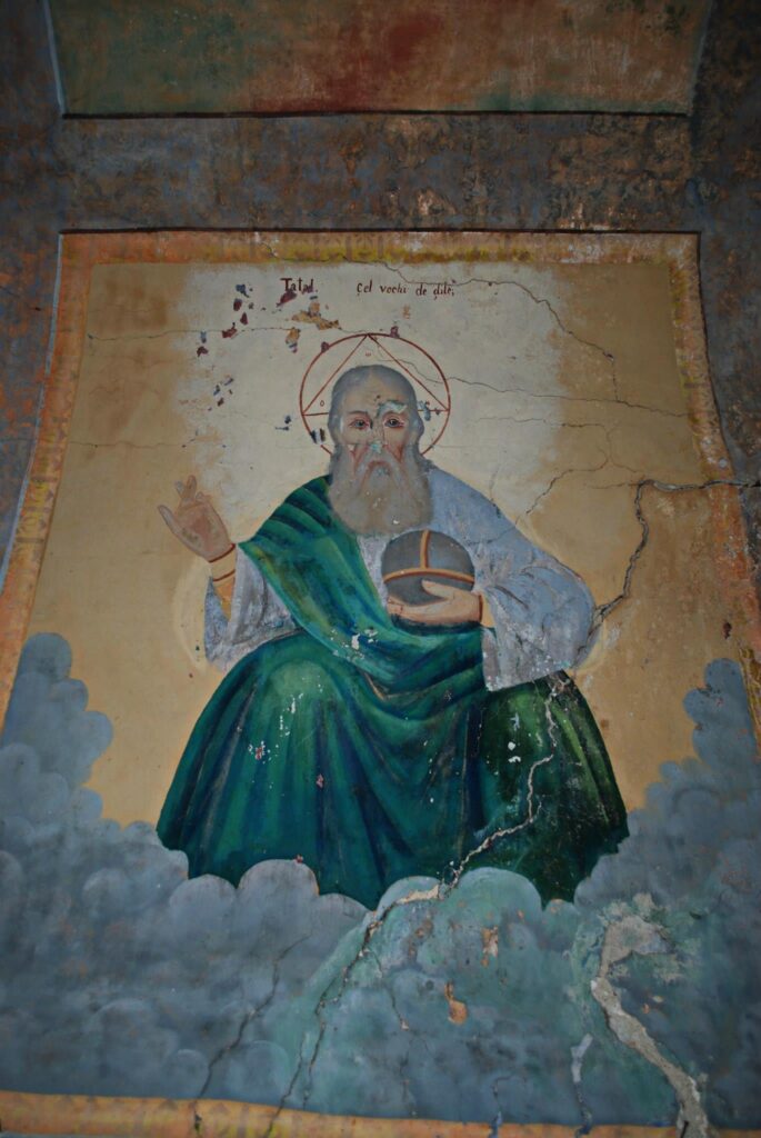 A weathered icon image of a saint with a halo, wearing a green robe and raising one hand in blessing; cracks and age marks are visible on the painting.