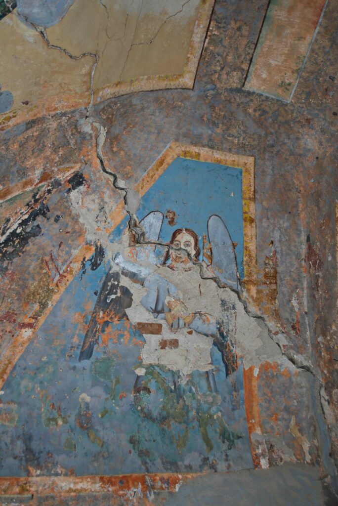 Faded wall painting with a blue panel showing part of an angel, heavily cracked and peeling plaster in orange, blue, and brown hues