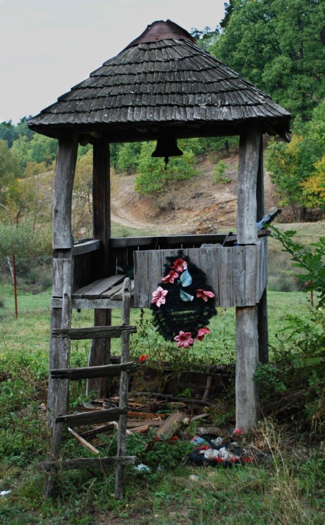 Weathered wooden bell tower on stilts in a grassy field, a bell hanging inside under a shingled roof, with a pink flower wreath on the front panel.