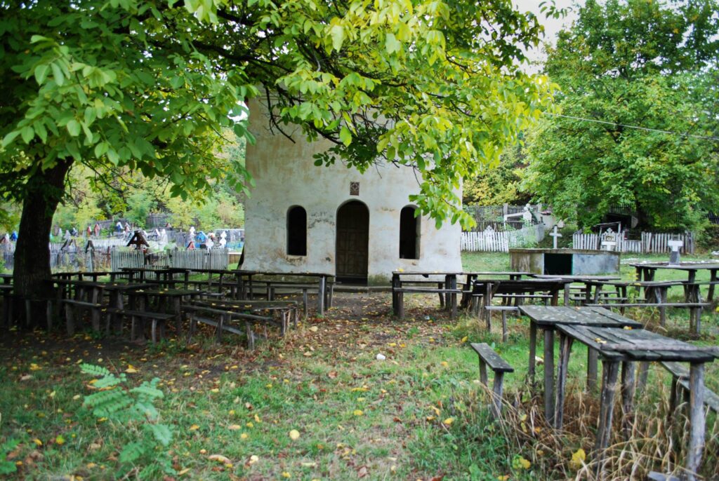 Whitewashed small chapel with three arched openings in a cemetery, surrounded by wooden benches and leafy trees.
