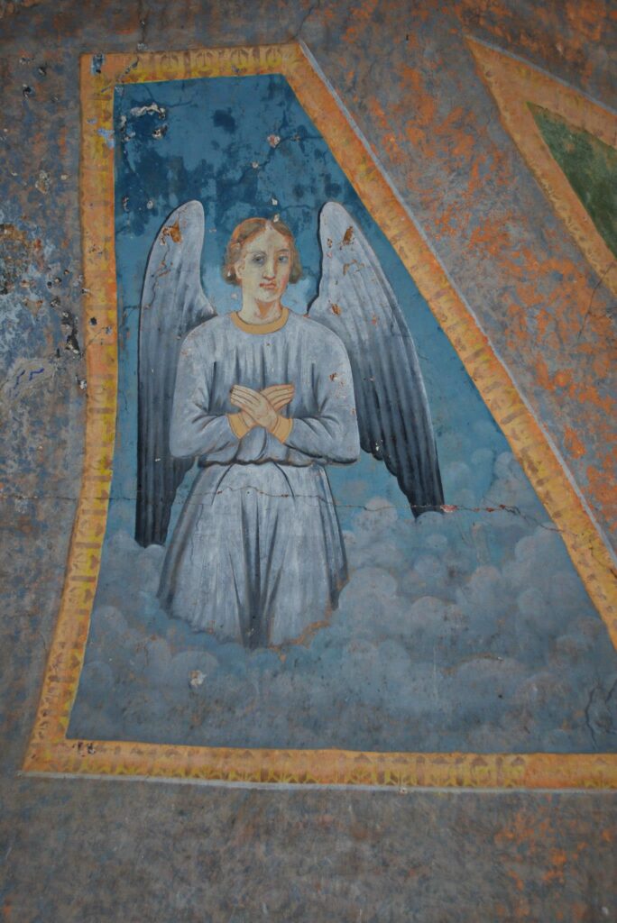 Worn mural of a pale angel with folded arms, white wings, and a blue robe framed by a blue panel and yellow-orange border on a weathered wall.