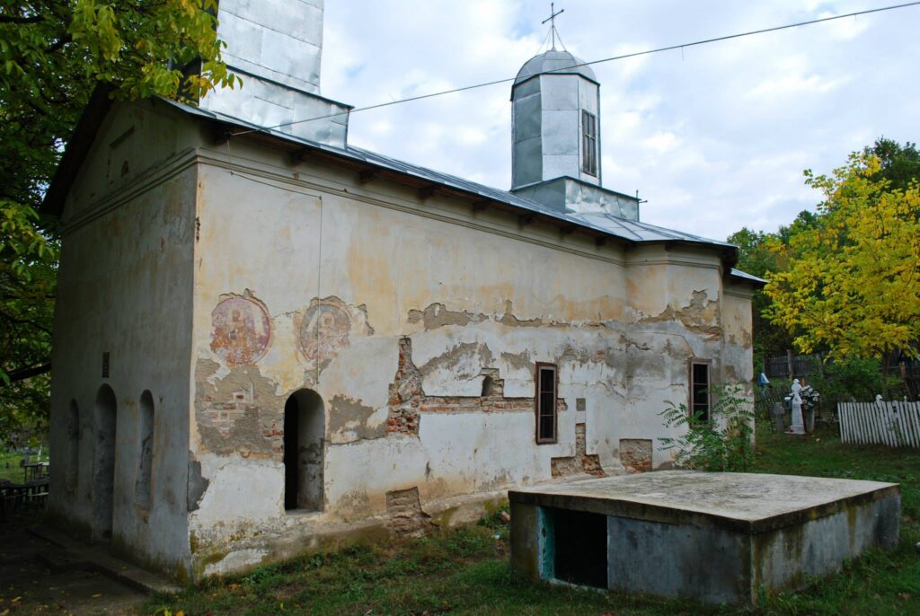 Old stucco church with worn walls, arched doorways, and fresco fragments, topped by a metal roof among green trees.