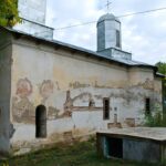 Old stucco church with worn walls, arched doorways, and fresco fragments, topped by a metal roof among green trees.
