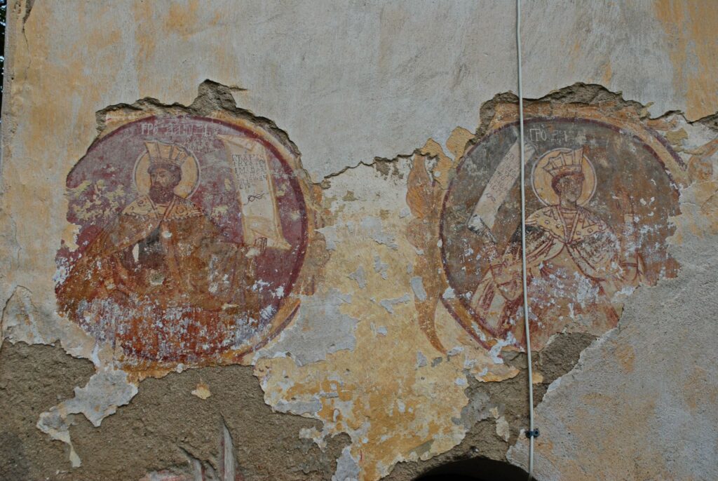Weathered wall with two faded circular frescoes of crowned figures, peeling plaster and a vertical pipe running down the center.