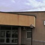 Exterior view of a beige public building with a glass entrance, two lamp posts, and a wheelchair ramp.