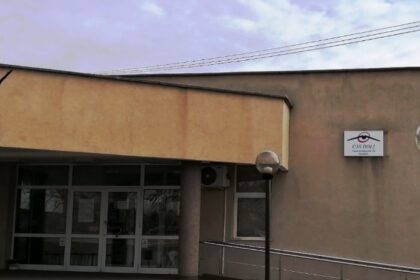 Exterior view of a beige public building with a glass entrance, two lamp posts, and a wheelchair ramp.