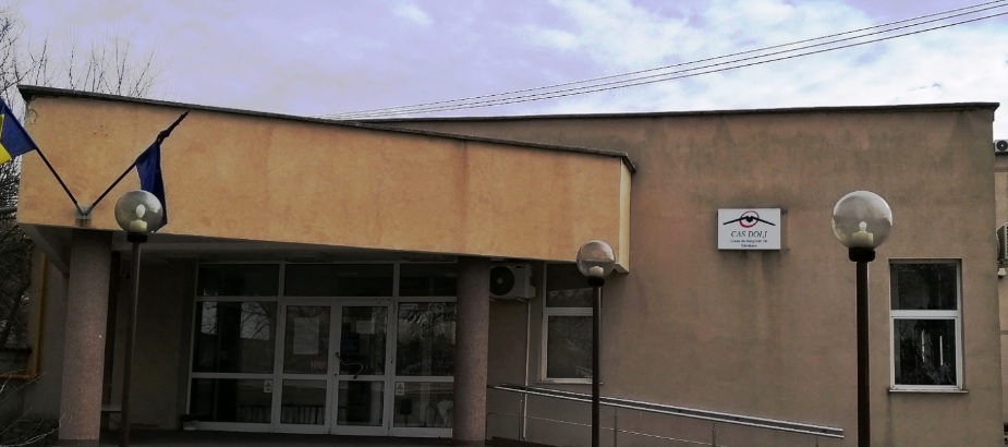 Exterior view of a beige public building with a glass entrance, two lamp posts, and a wheelchair ramp.
